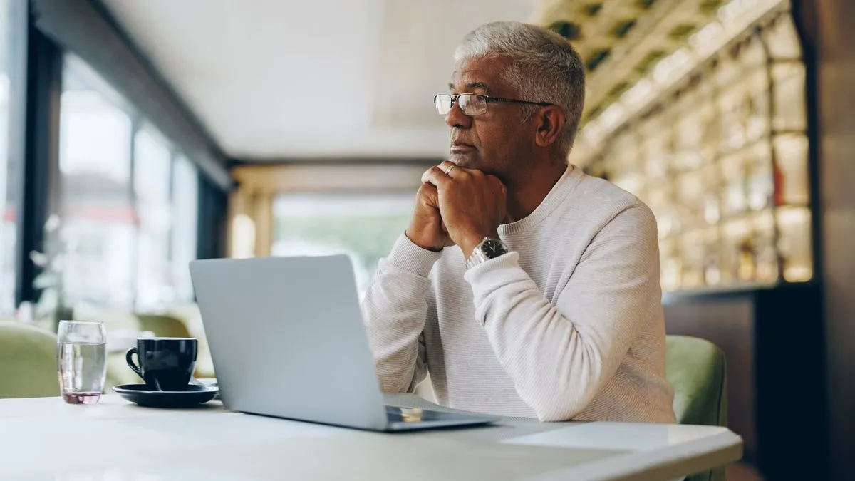 Mature man in a light sweater sitting at a table with a laptop, coffee cup, and glass of water in a bright café.