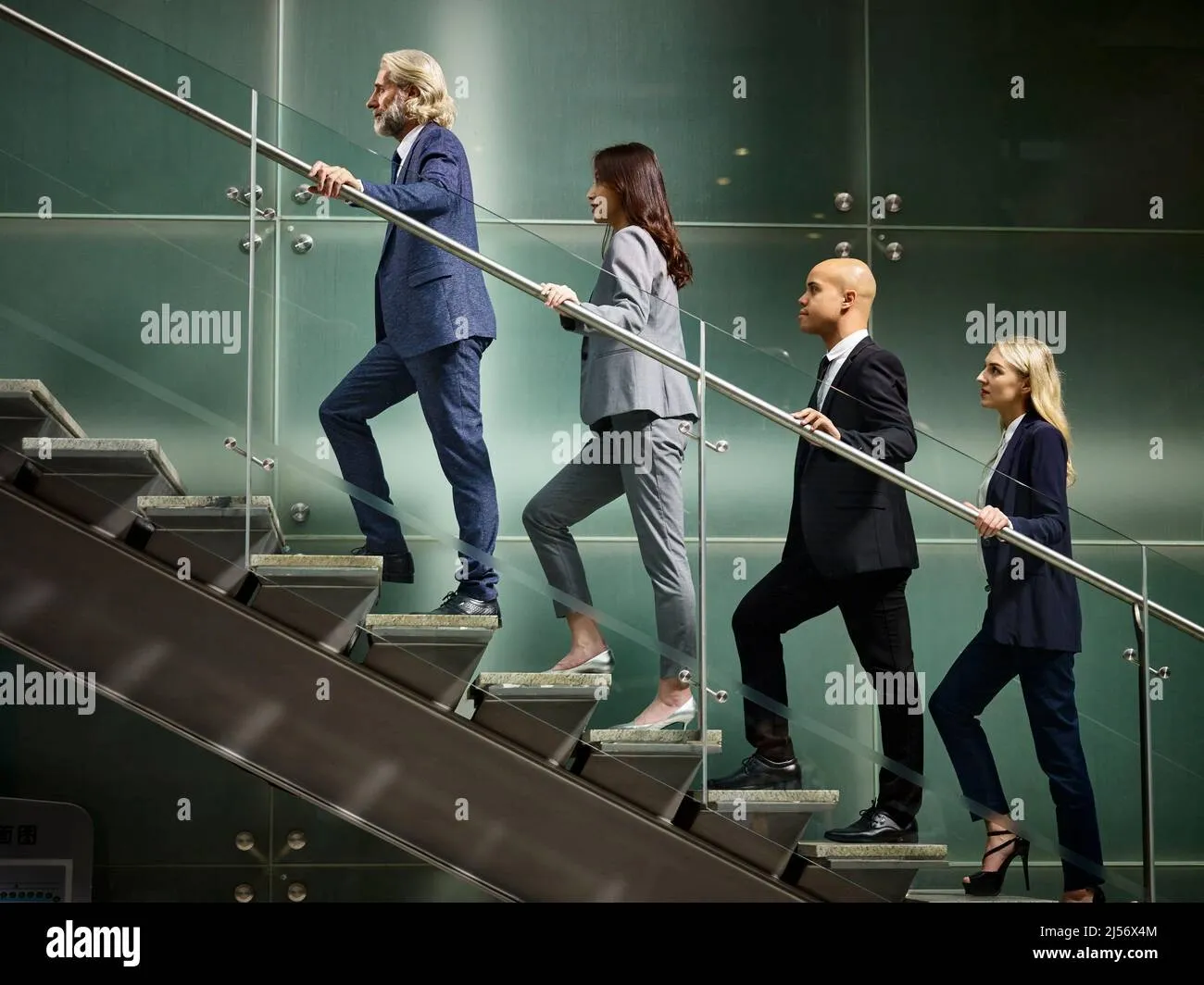 Four business professionals dressed in suits ascending a modern glass staircase inside an office building.