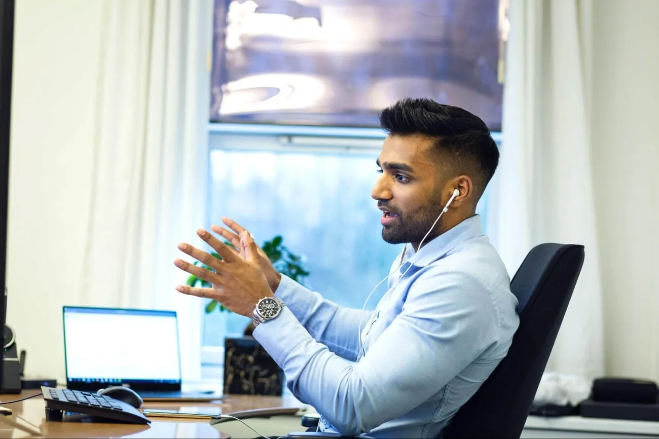 Man in a light blue shirt wearing earphones gestures while sitting at a desk with a laptop in a bright office.
