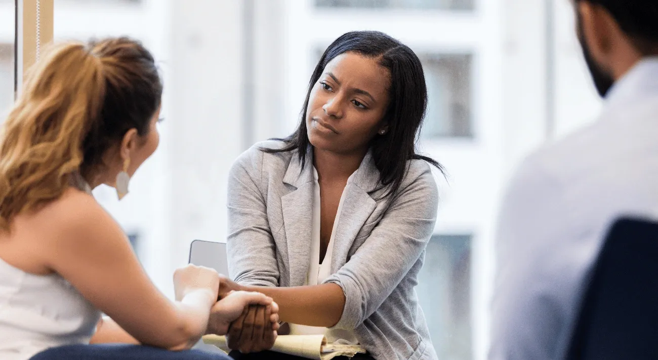 Businesswoman in gray blazer comforting a seated colleague by holding her hands during a meeting in an office.