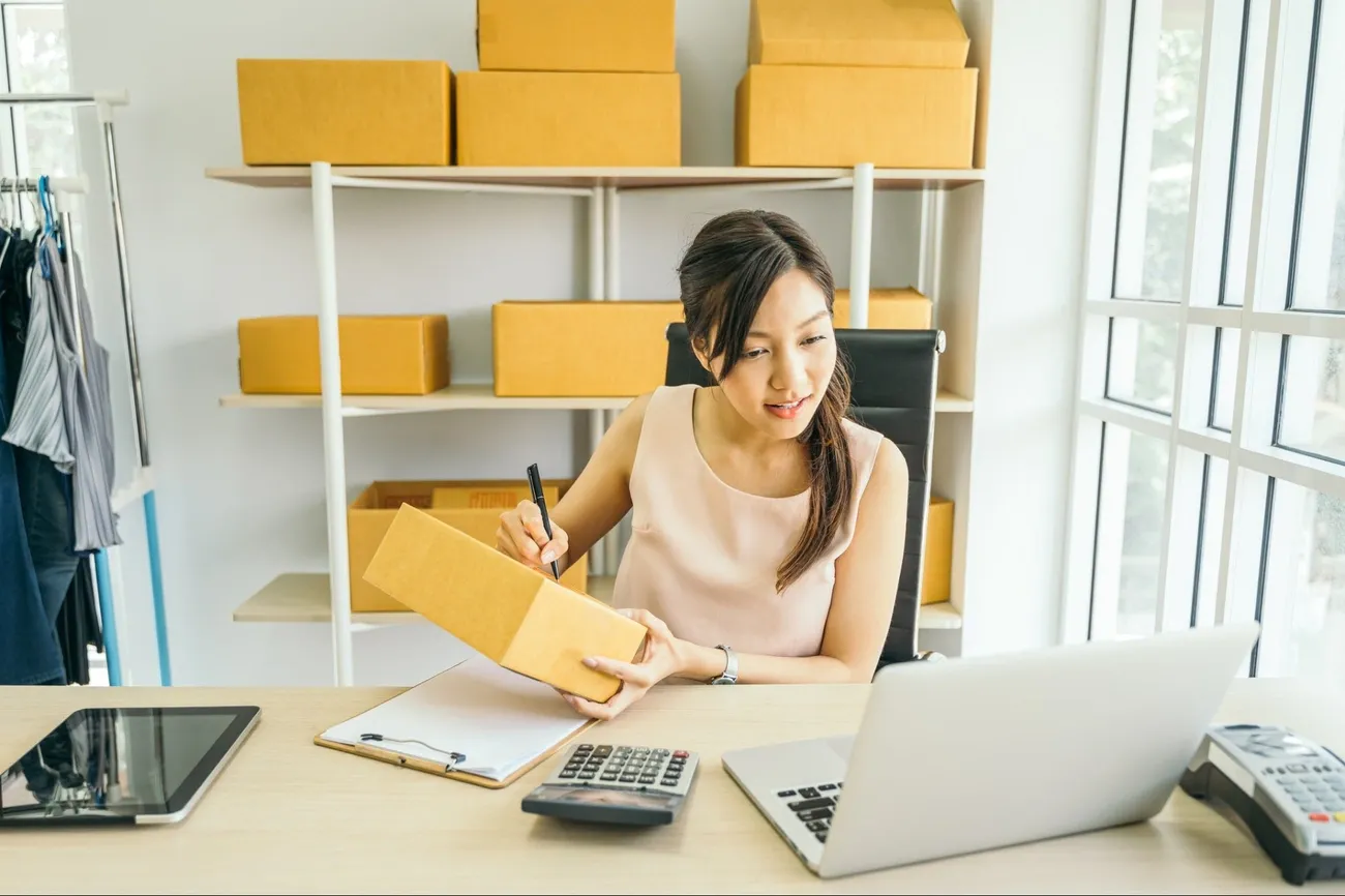 Person labeling a cardboard box at a desk with laptop, calculator, clipboard, and shelves of boxes in the background.